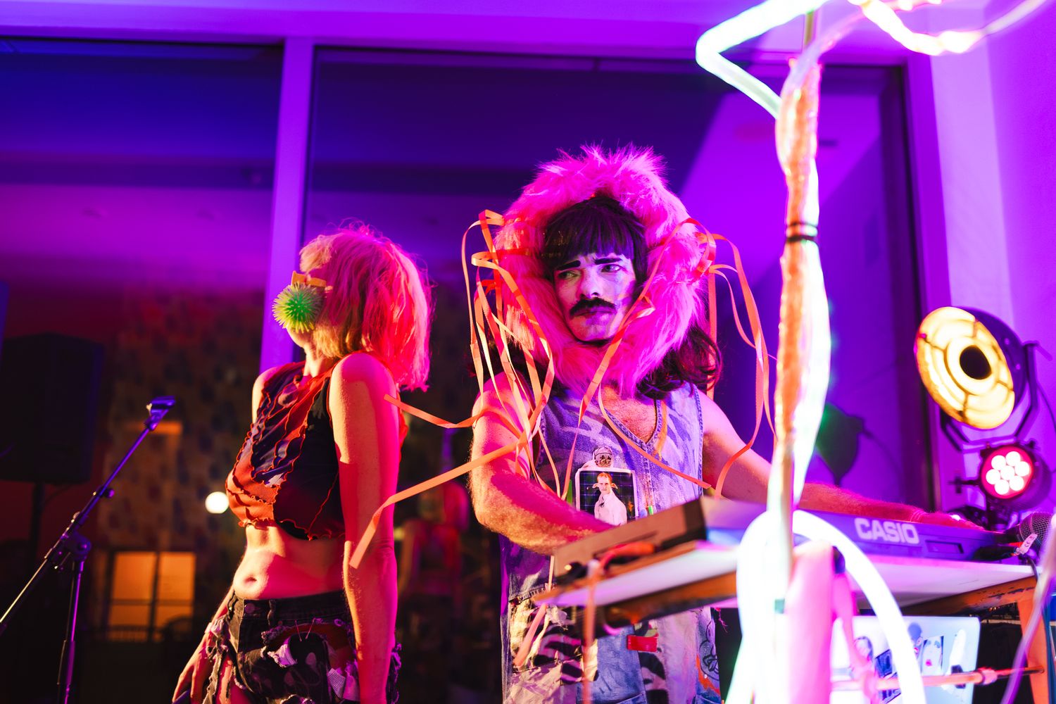 Two colorful performers on stage with neon lights, featuring wild pink hair and funky outfits at a vibrant music event.