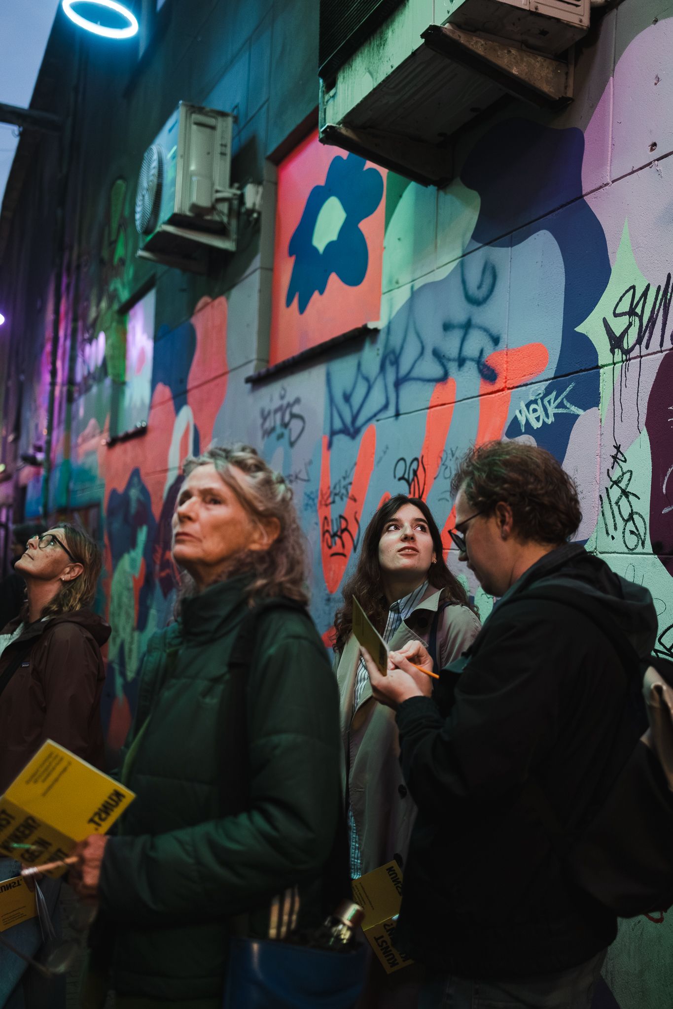 People admire colorful graffiti murals on a city alley wall at night, with vibrant street art illuminated behind them.