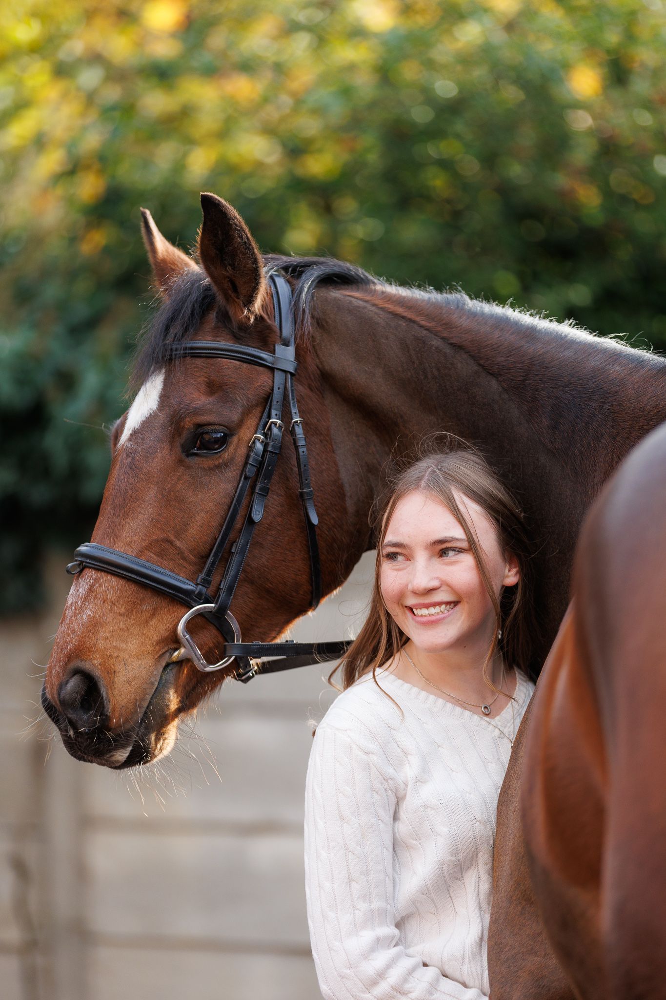 A beautiful bay horse wearing a black bridle stands next to someone in a white lace dress in an outdoor setting.