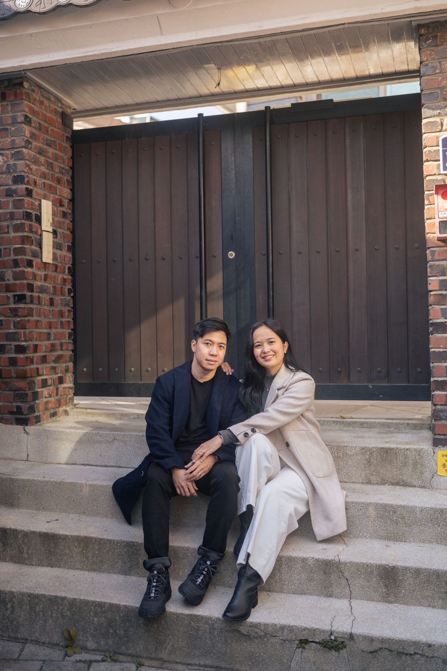 A couple sits together on concrete steps outside a building with dark wooden doors and brick walls.
