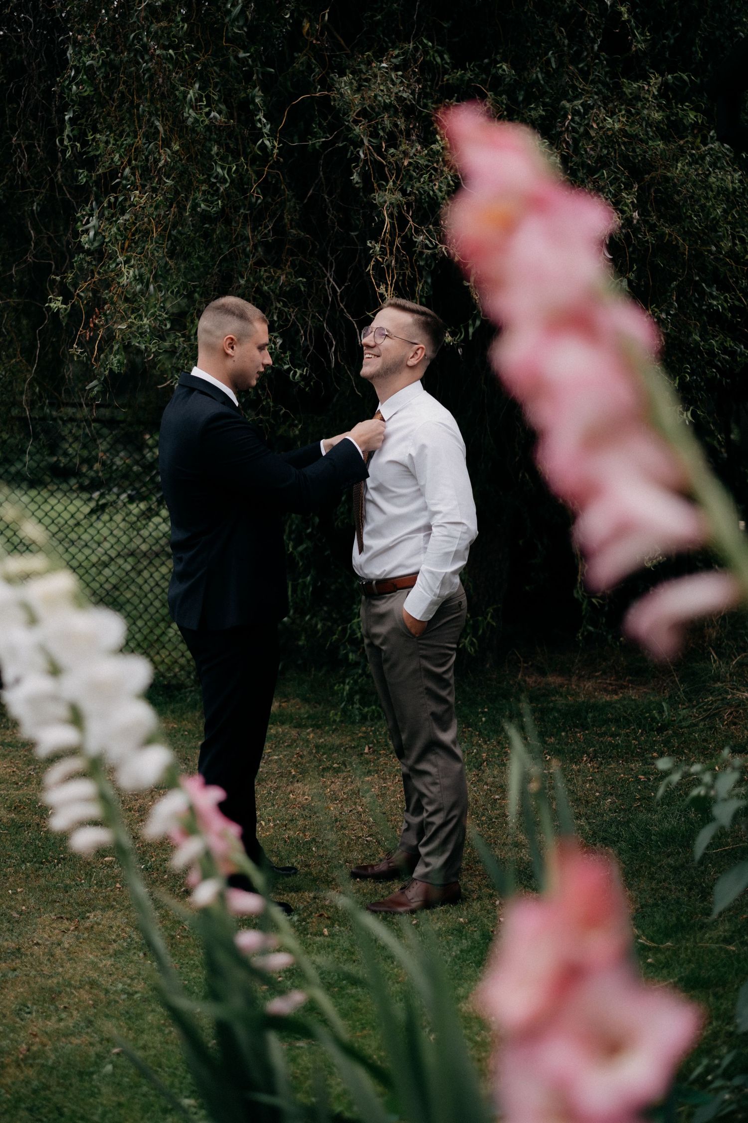Two people in formal wear share a moment in a garden with pink flowers in the foreground.
