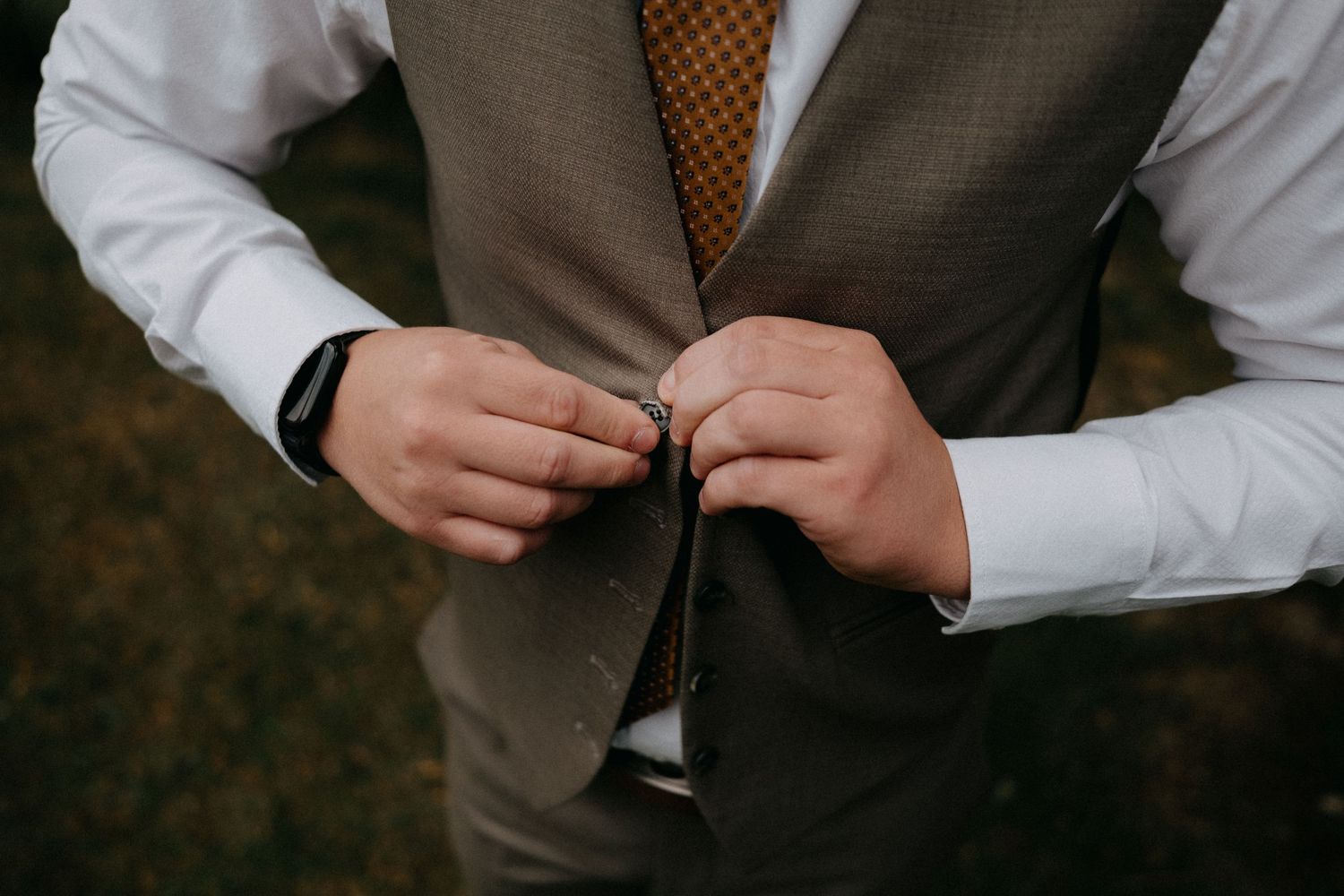 Close-up of hands buttoning a brown vest over a white dress shirt.