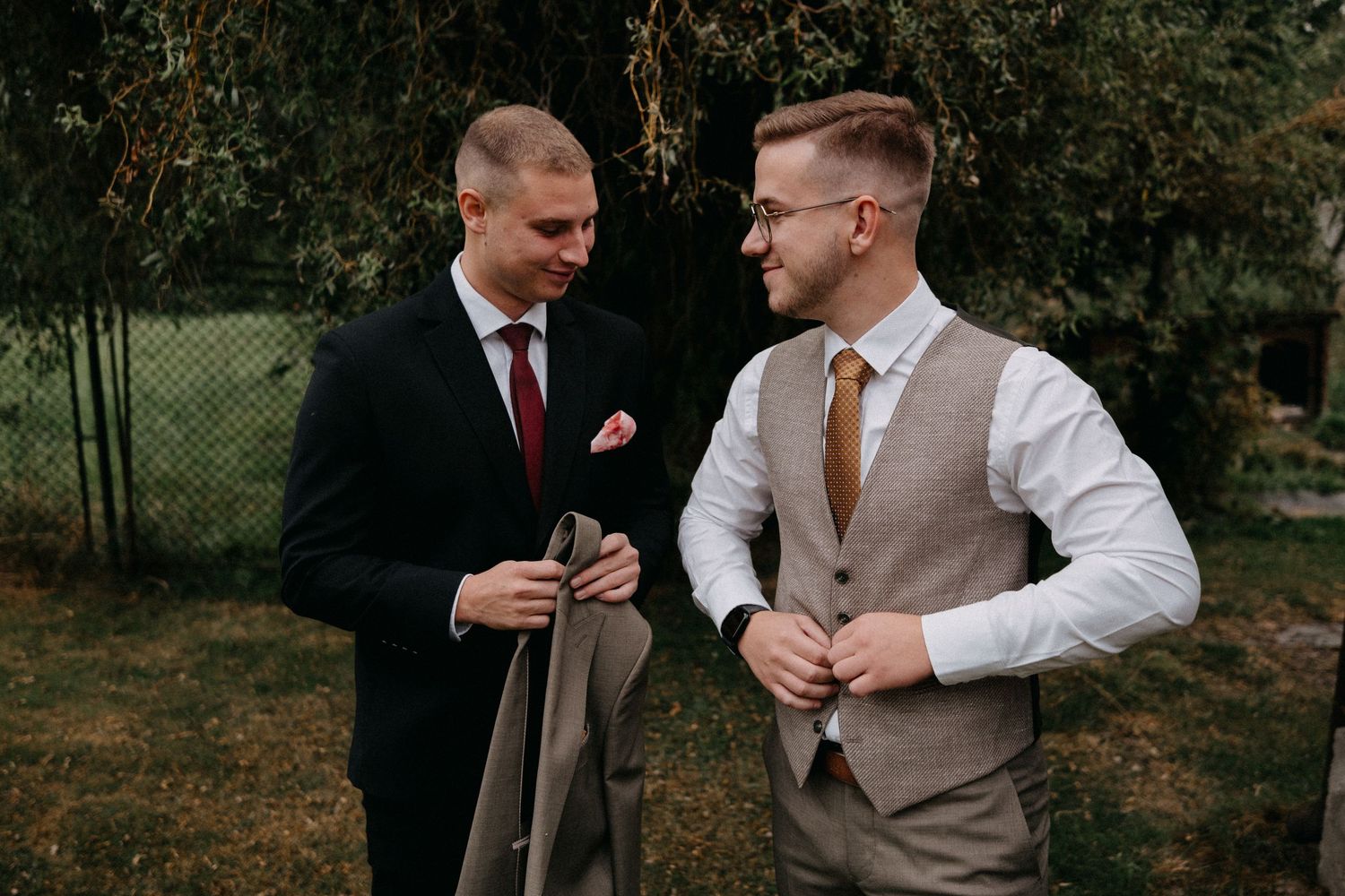 Two people in formal attire conversing outdoors near garden foliage.