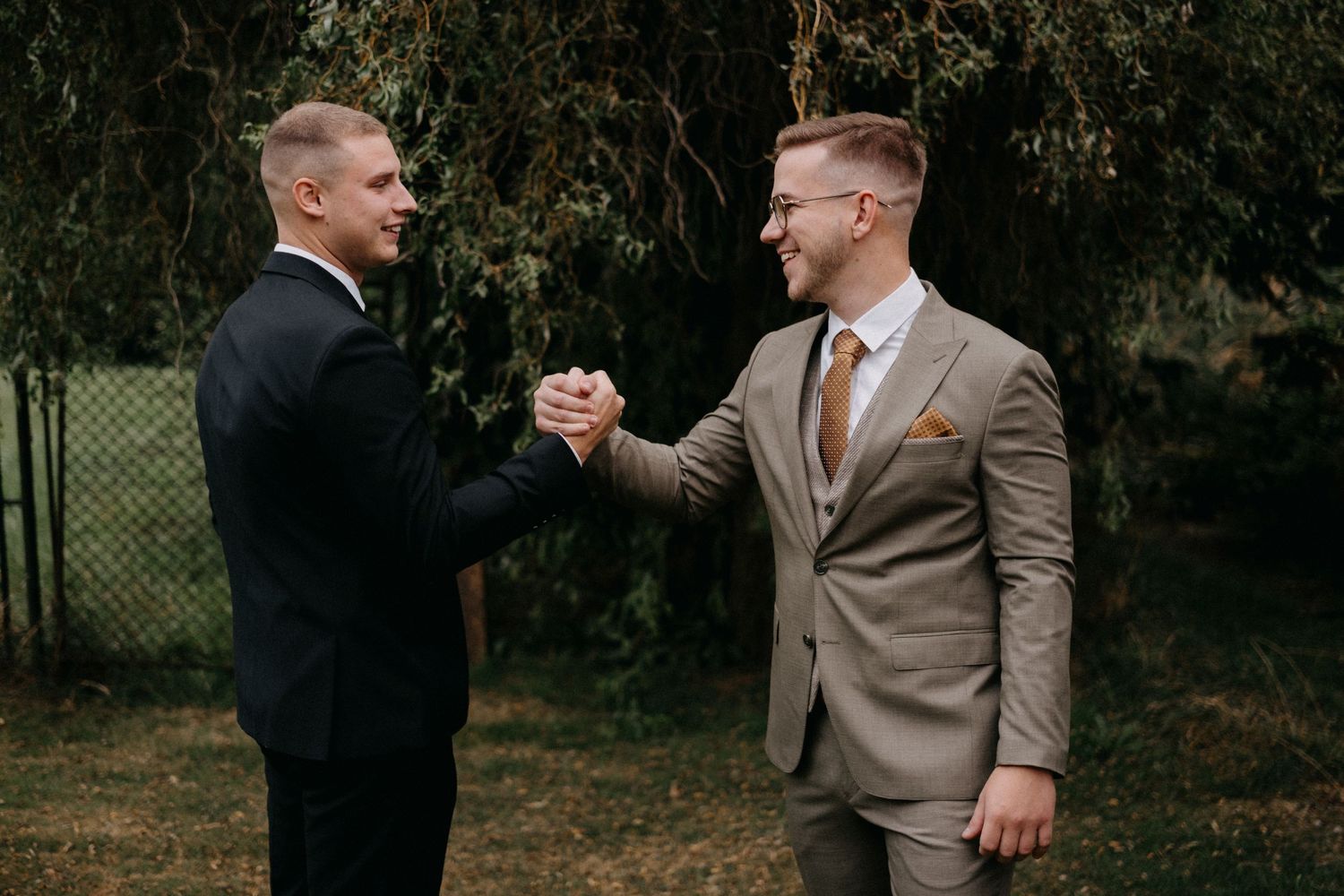 Two people in suits shake hands in an outdoor setting.