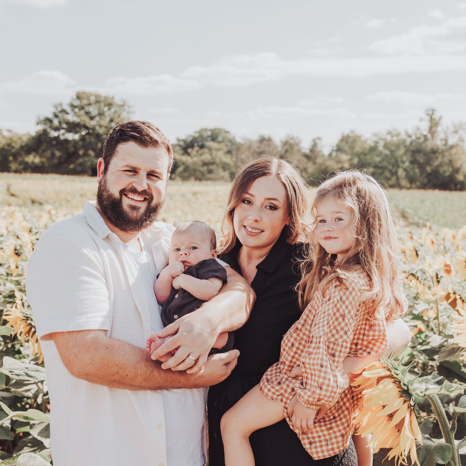 Family of 4 in a sunflower field.  The dad is holding their newborn baby boy and the mum is holding their older daughter.