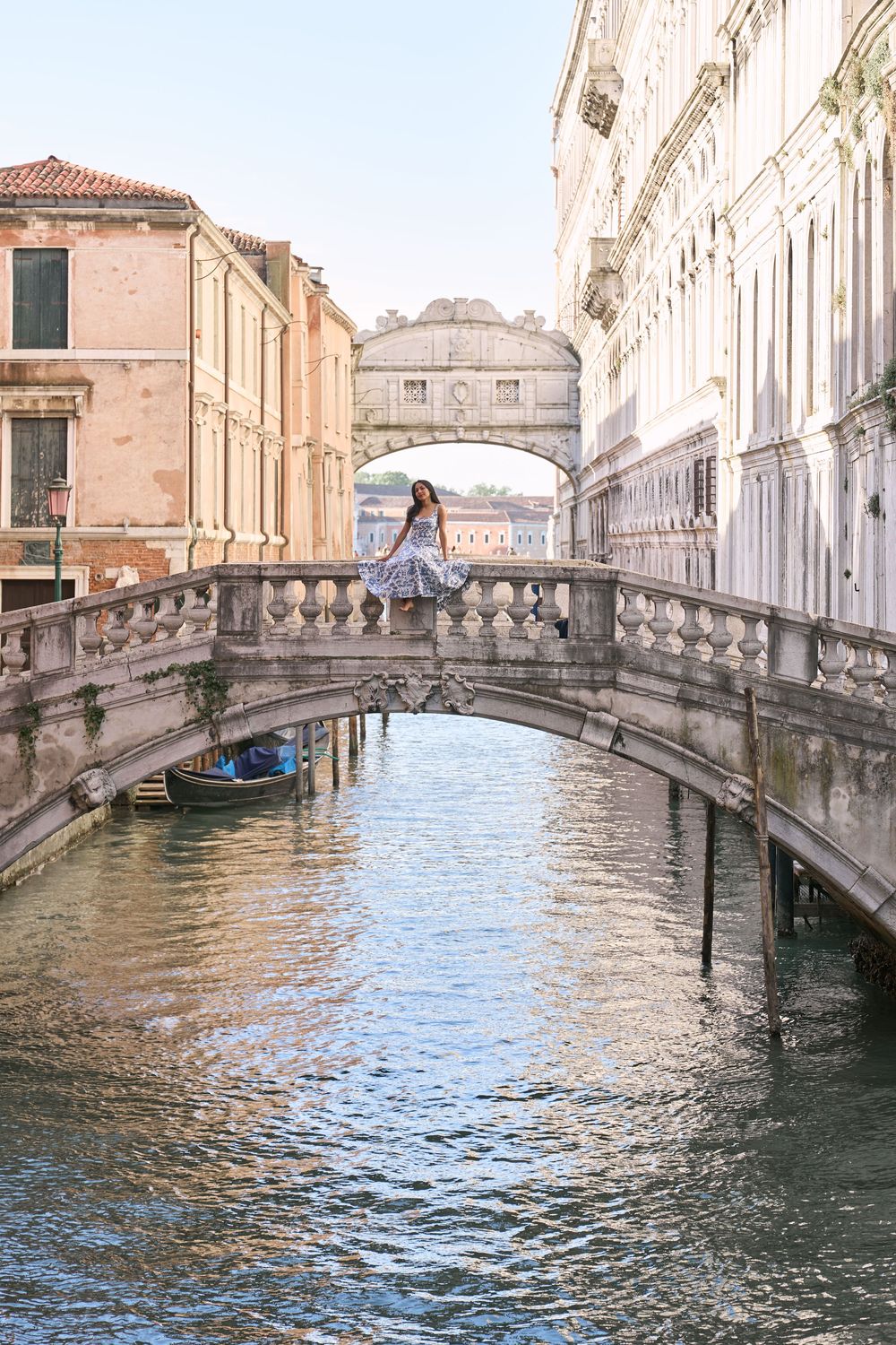 Woman in floral dress sitting on ornate stone bridge over Venice canal with Bridge of Sighs in background.