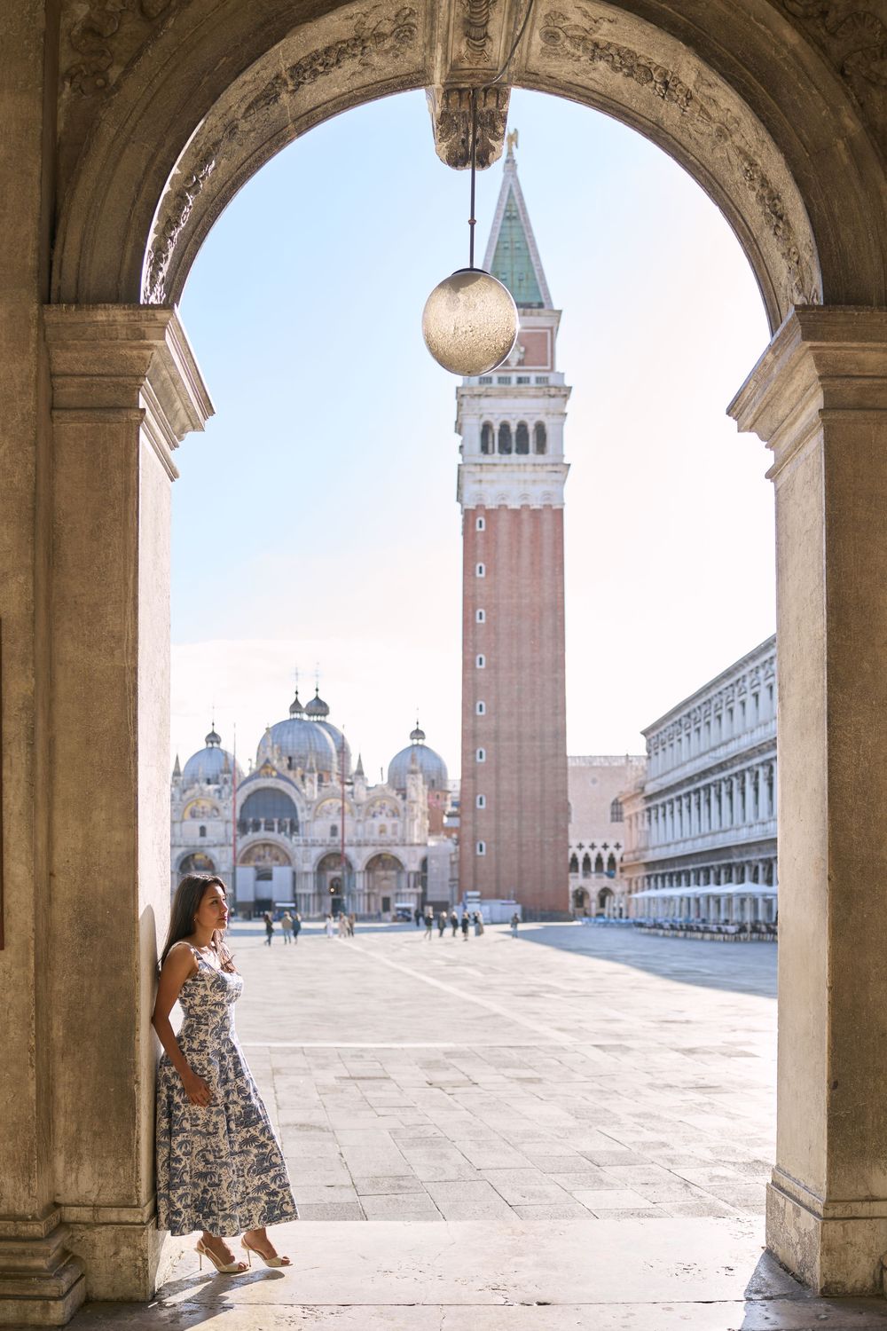 Woman in floral dress posing under an arch in St. Mark's Square, Venice, with the Campanile bell tower.