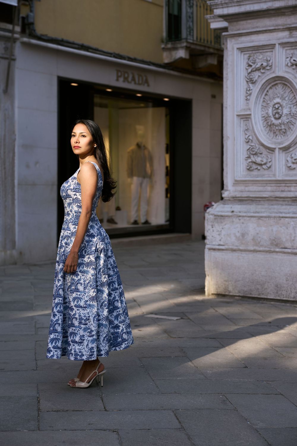 Woman in blue floral midi dress posing on cobblestone street outside Prada store in Venice, Italy.