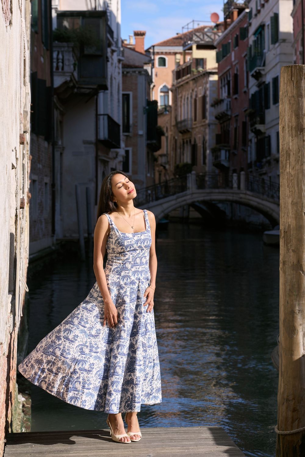 Woman in blue floral midi dress posing on wooden dock beside Venice canal with bridge and historic buildings.