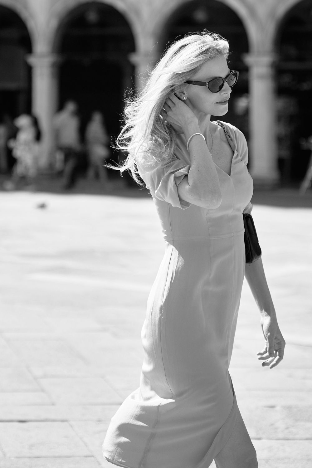 Blonde woman in white dress and sunglasses walking through a sunny European plaza in black and white.