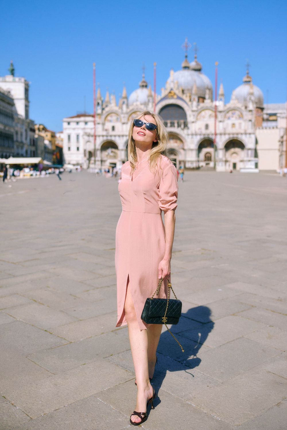 Blonde woman in pink midi dress with black handbag posing in St. Mark's Square, Venice, Italy.
