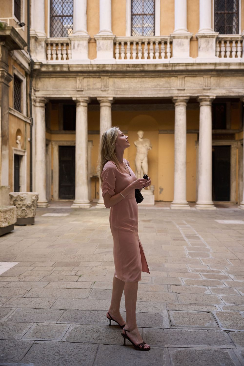 Blonde woman in pink dress gazing upward in a classical European courtyard with columns and stone architecture.