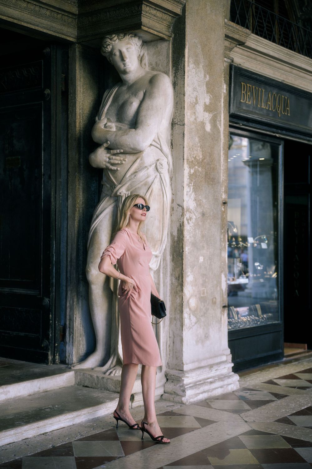 Blonde woman in pink midi dress poses by classical statue outside luxury Bevilacqua store in Venice.