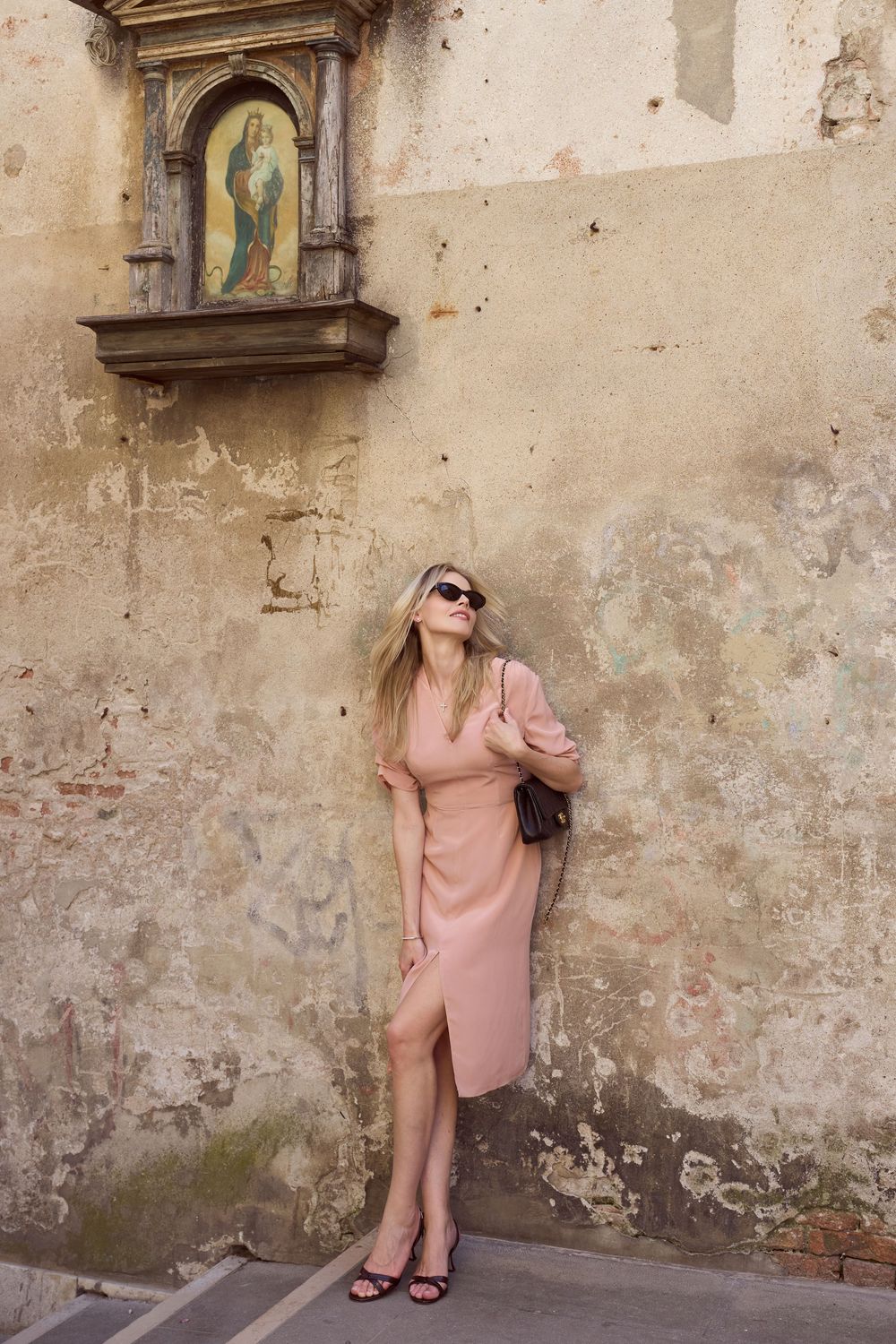 Woman in pink dress and sunglasses leaning against aged wall with religious icon niche in European city.