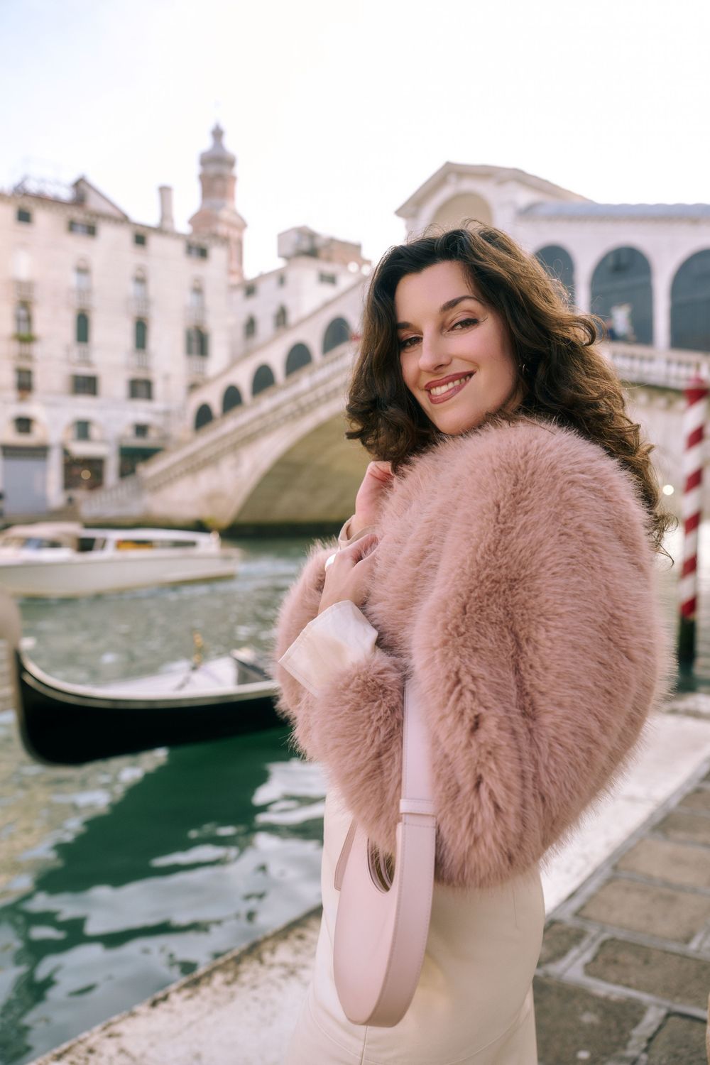 Woman in pink fur coat poses along Venice canal with historic architecture and gondolas in background. Book your Venice photoshoot.