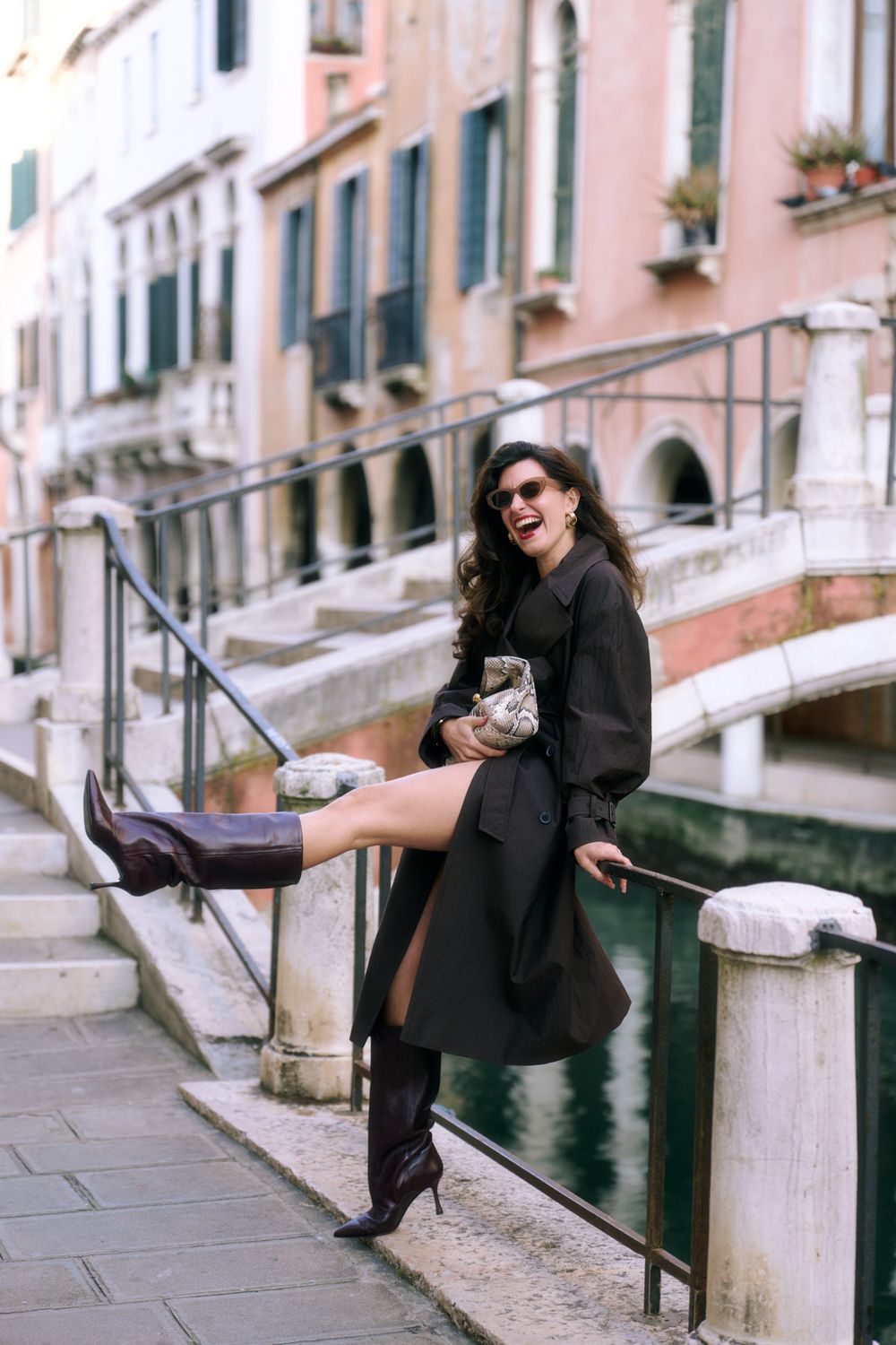 Woman in black coat and boots sits playfully on railing along a charming European street with pink buildings.