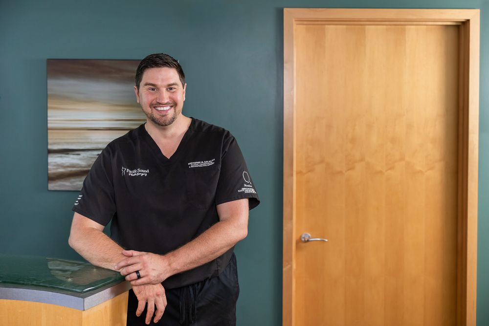 Plastic surgeon in Bellevue, WA male practitioner lifestyle headshot wearing black scrubs standing in office with arm on countertop