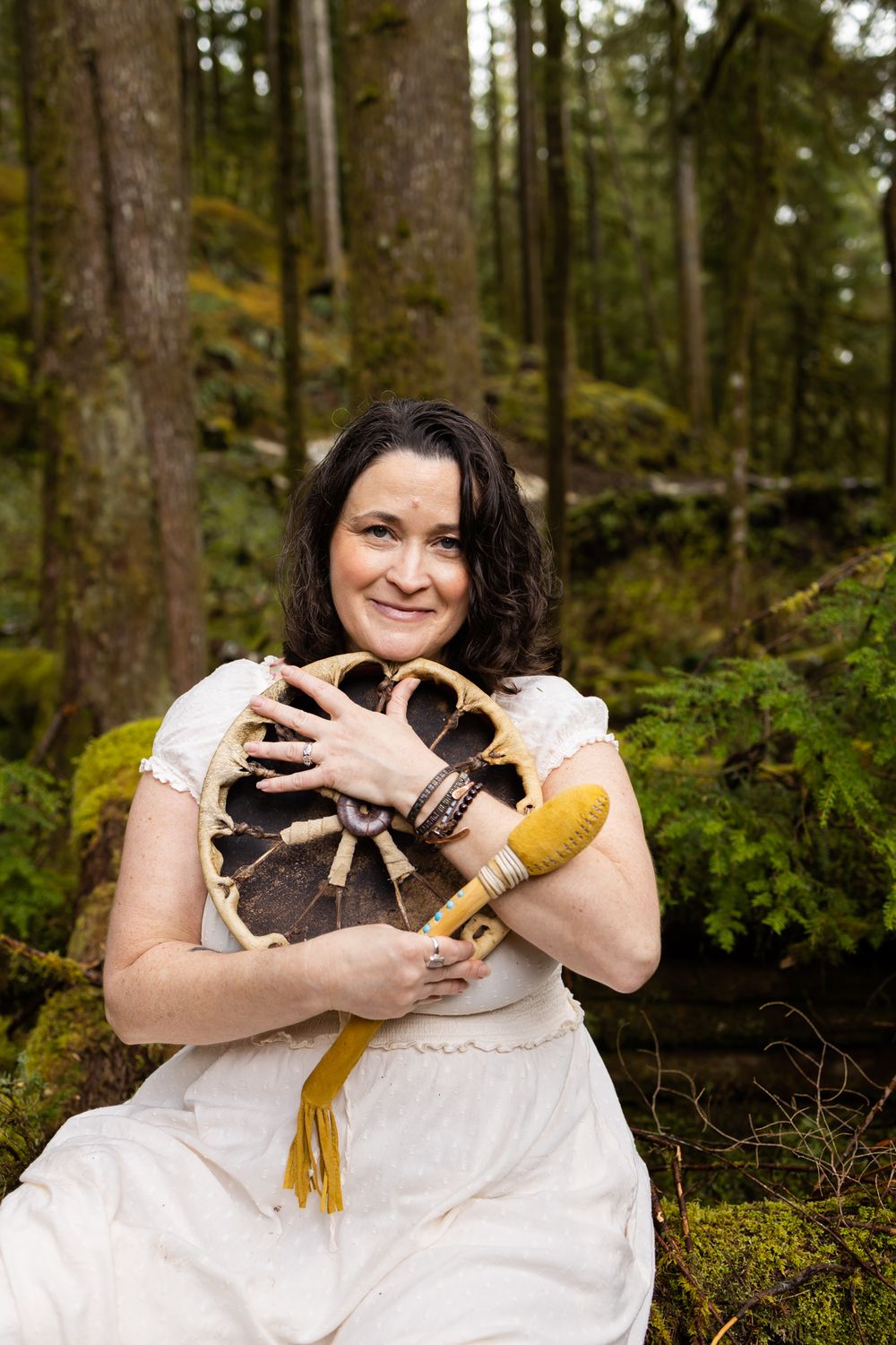Kerry drum maker and energy worker hugging drum in forest smiling at camera in Sammamish, WA