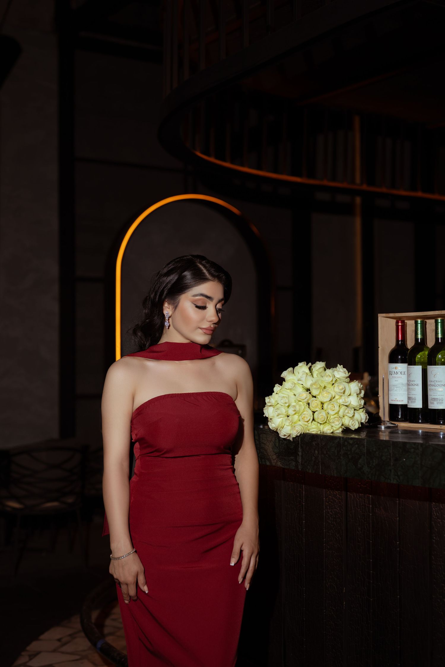 Woman in burgundy evening dress poses elegantly against dark bar setting with white roses in background.