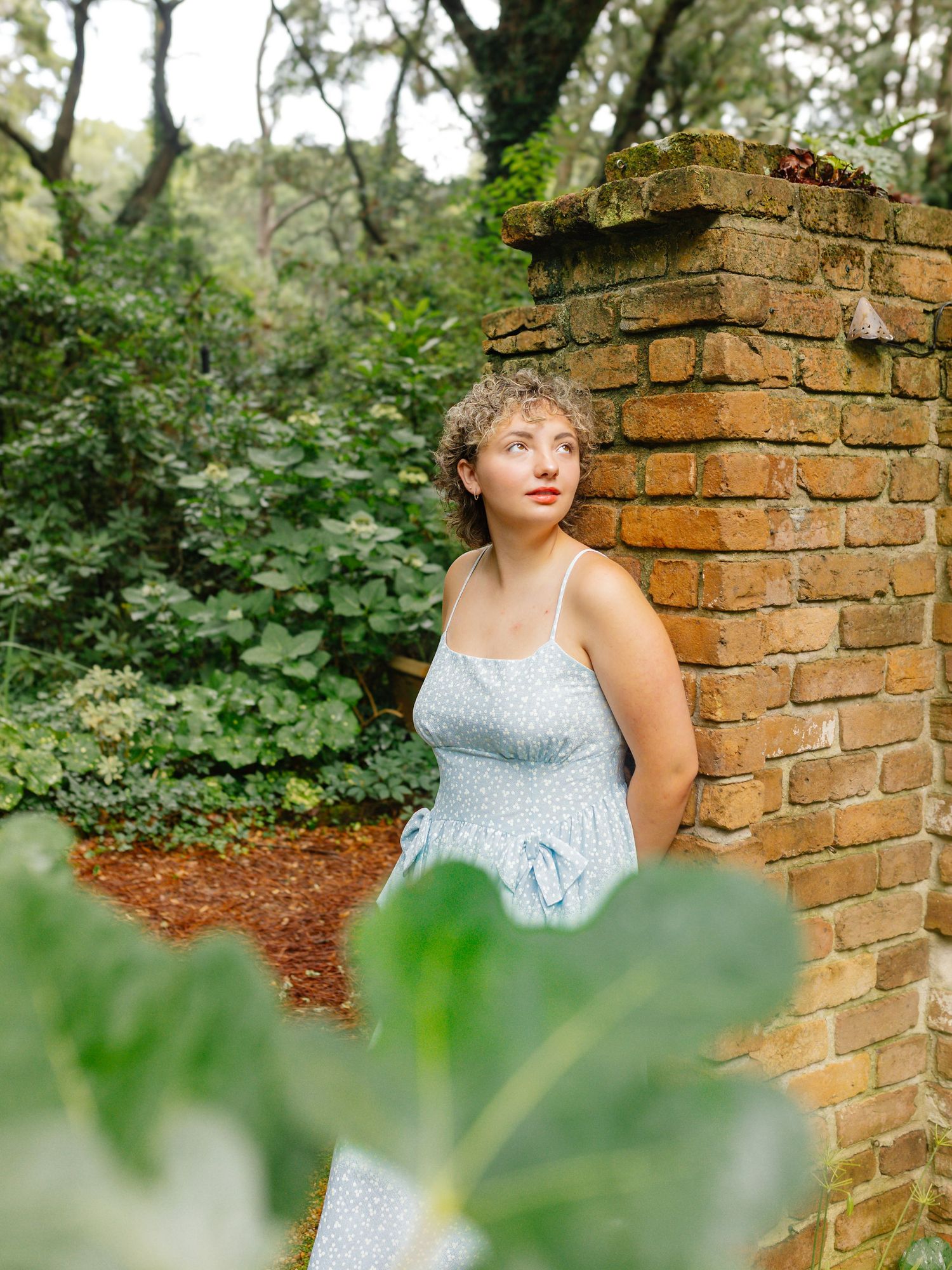Woman in blue dress poses against rustic brick wall surrounded by lush greenery.