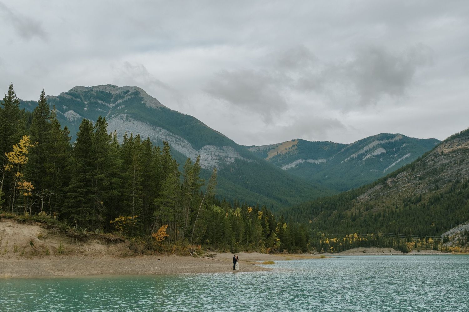 A scenic mountain landscape with pine trees and a tranquil lake under cloudy skies in the Canadian Rockies.