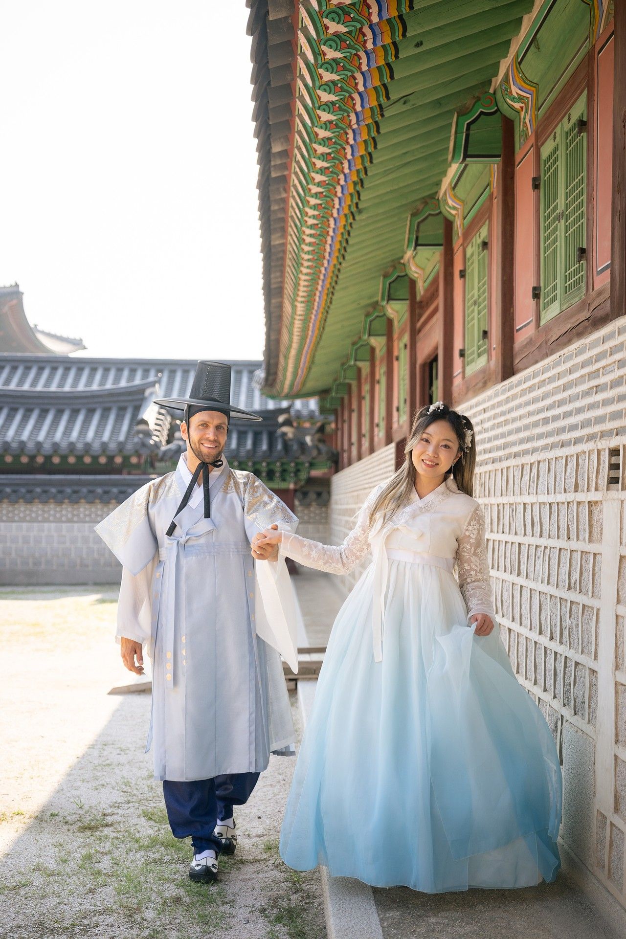 A couple in traditional Korean hanbok clothing poses along a temple corridor with ornate green and red painted beams.