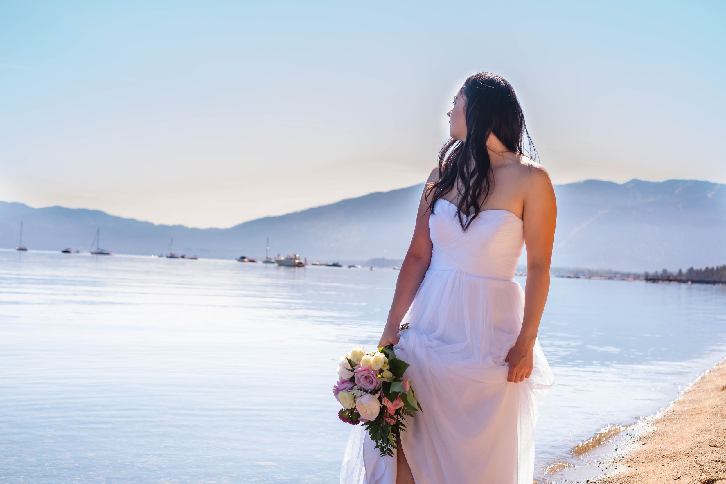 A person in a white wedding dress walks along a the crystal clear shores of lake tahoe in california.