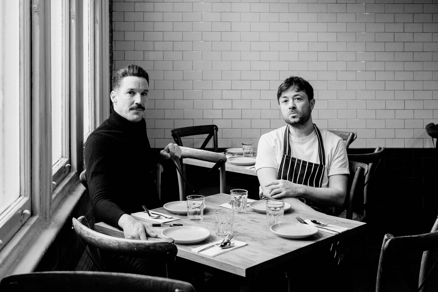 Two people sit at a restaurant table near a window in a black and white photograph.