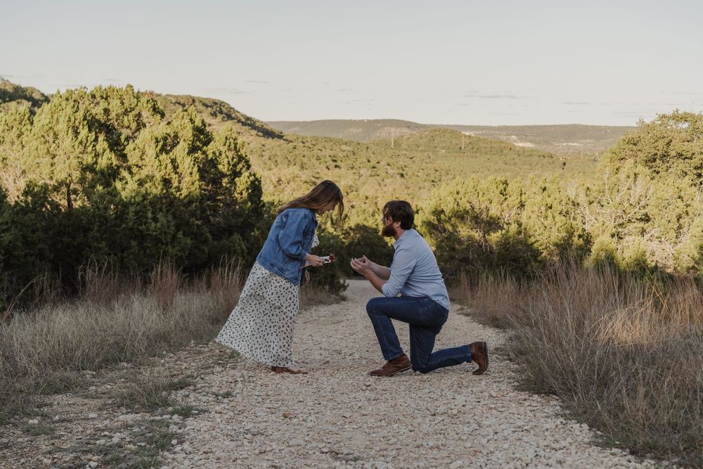 A couple shares an intimate moment on a dirt path surrounded by desert brush during a sunset proposal.