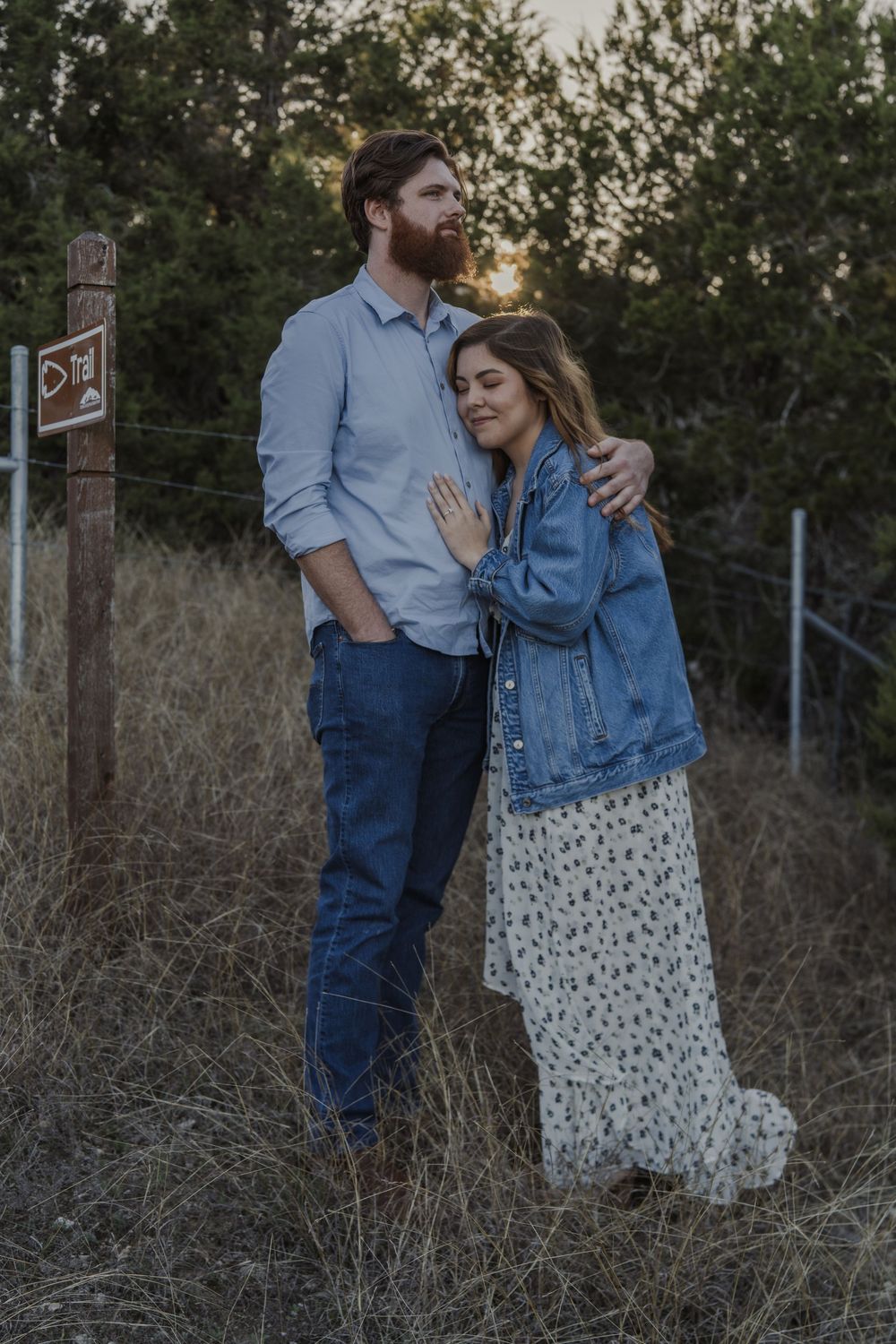 A couple embraces on a nature trail during golden hour while wearing casual attire.
