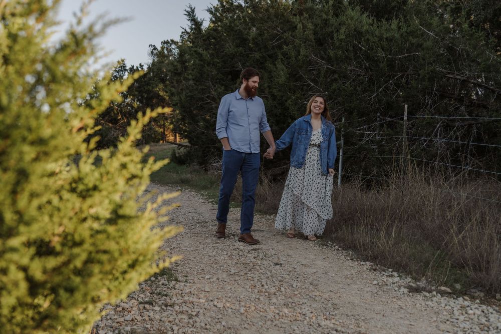 A couple walks hand in hand on a gravel path surrounded by green foliage at dusk.