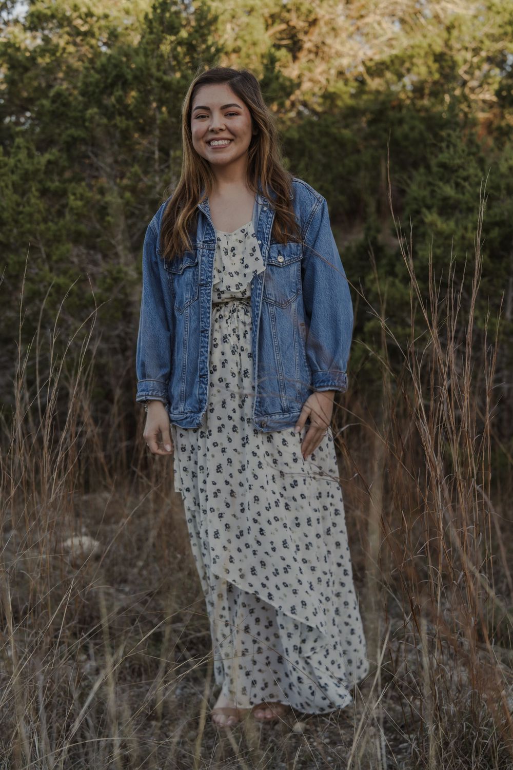 A woman in a floral dress and denim jacket stands smiling in a natural outdoor setting.
