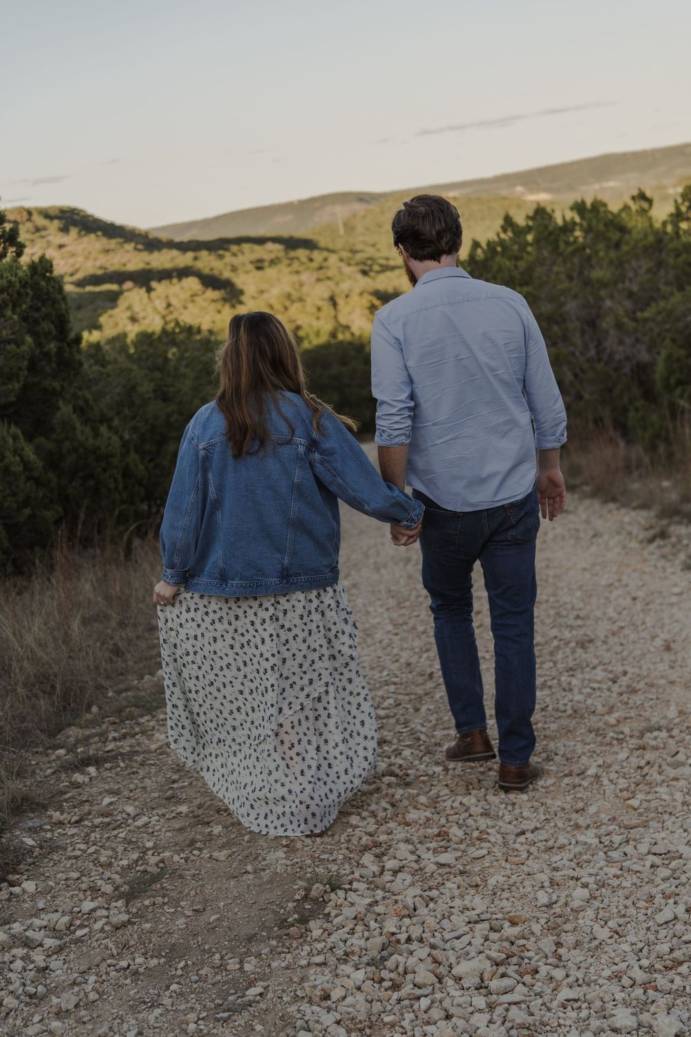 A couple walks hand in hand on a rocky path through desert brush at sunset.