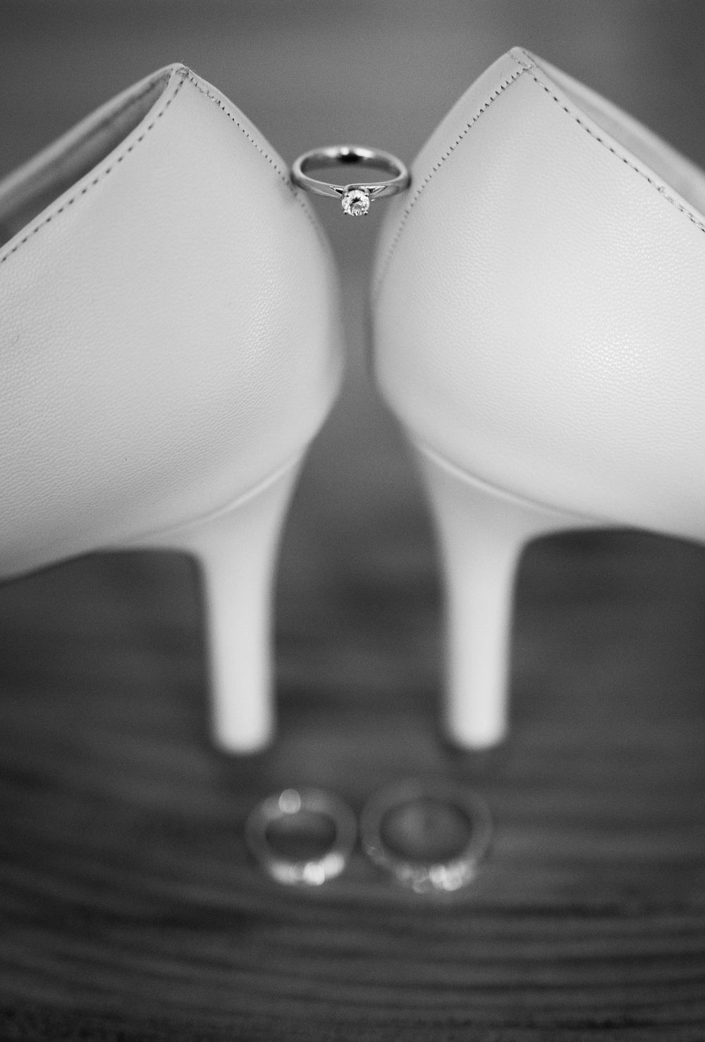 Wedding rings rest beside elegant white bridal shoes on a wooden surface in black and white.