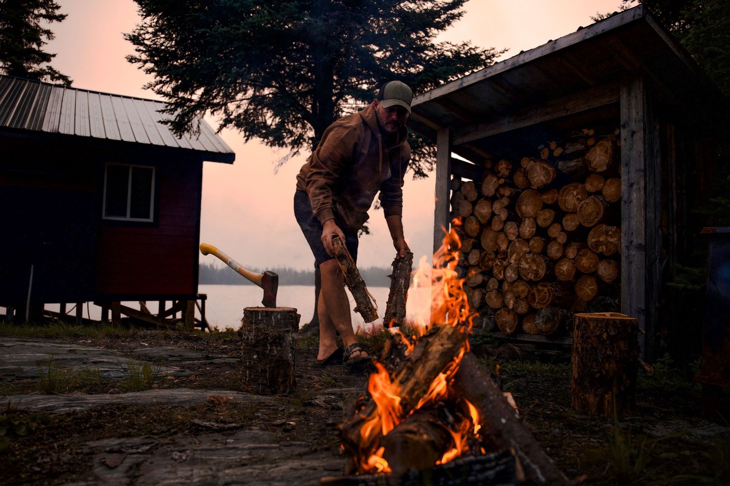 A rustic cabin with stacked firewood and a campfire at sunset.