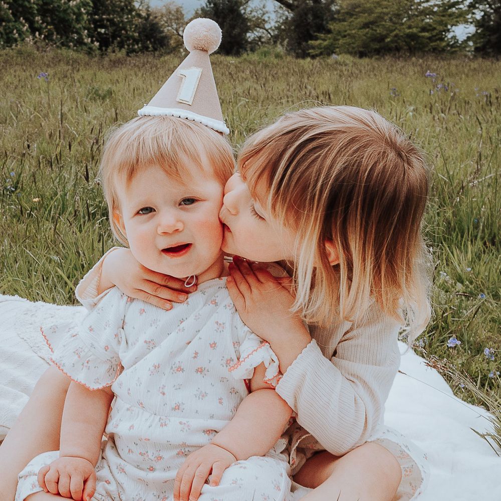 Fun image of girl wearing a number one hat to celebrate first birthday.  Her sister is sitting behind her giving her a kiss on the cheek.