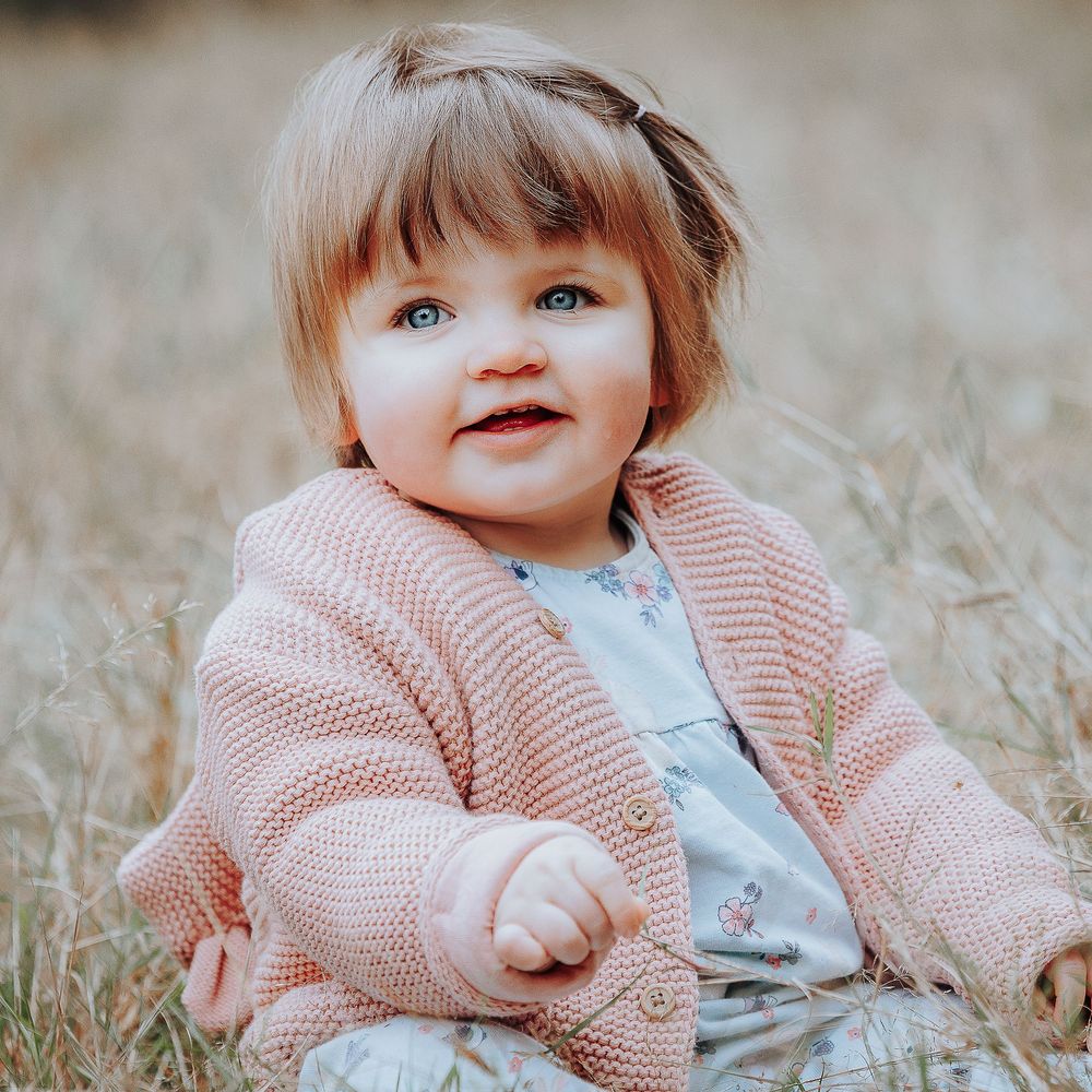 Baby girl sitting in a meadow smiling with a navy blue bow in her hair and a pink cardigan.