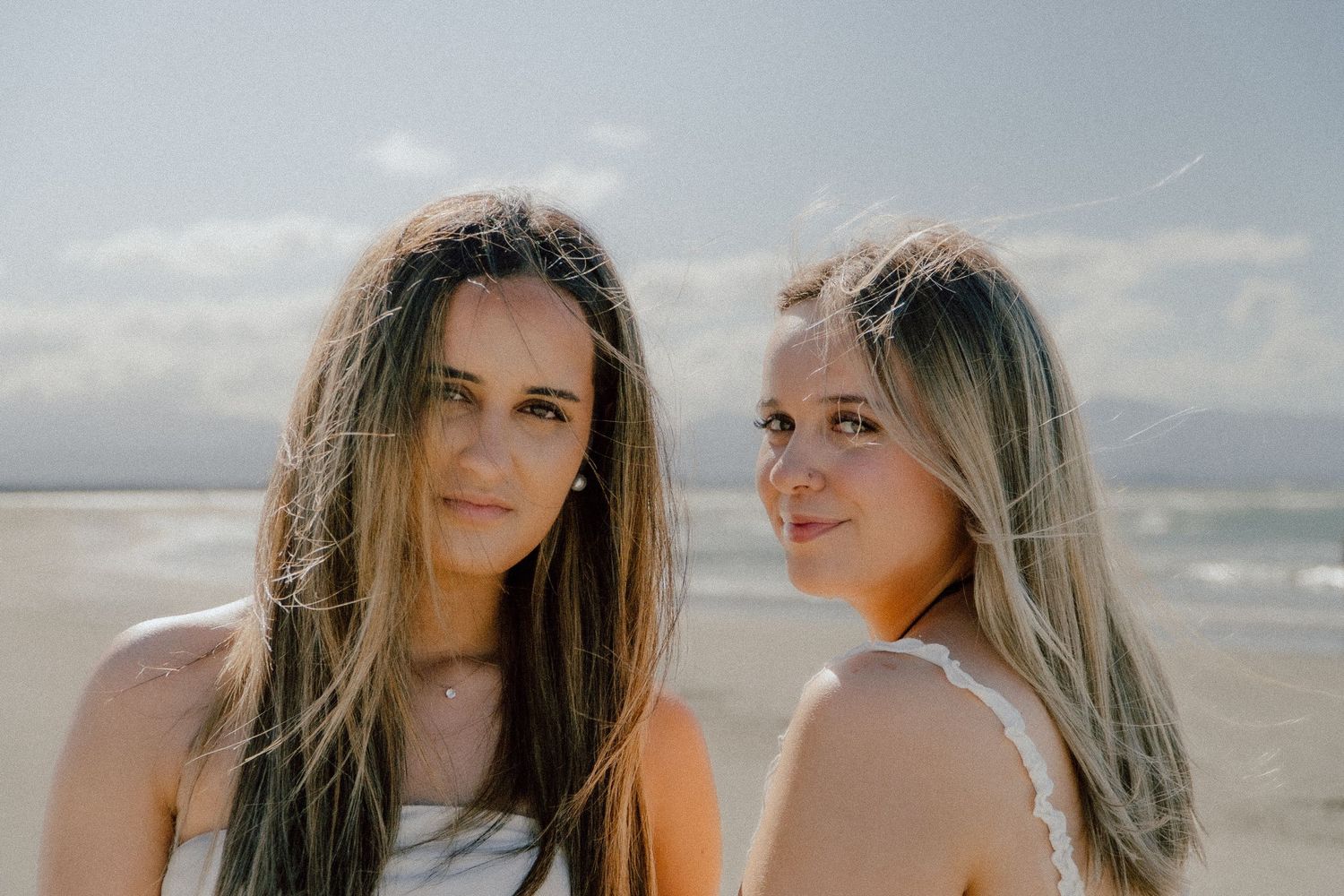 Two people with long hair smile at the beach on a sunny day with ocean waves in the background.