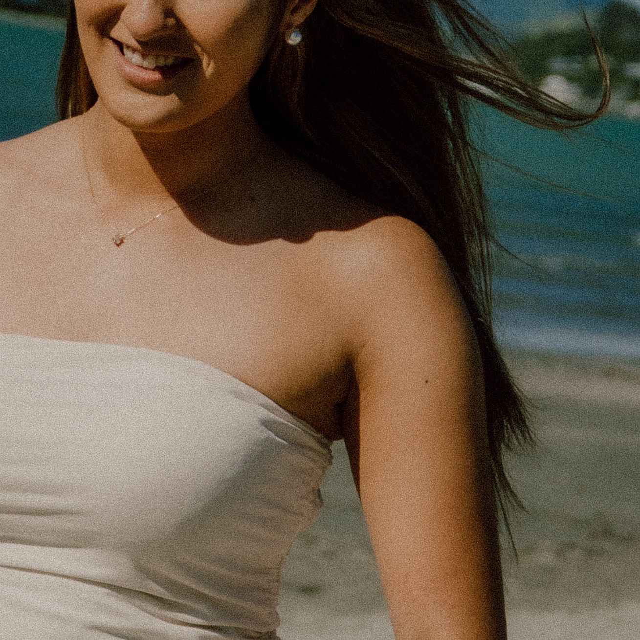 A person in a white strapless top smiles at the beach with dark hair flowing in the wind.