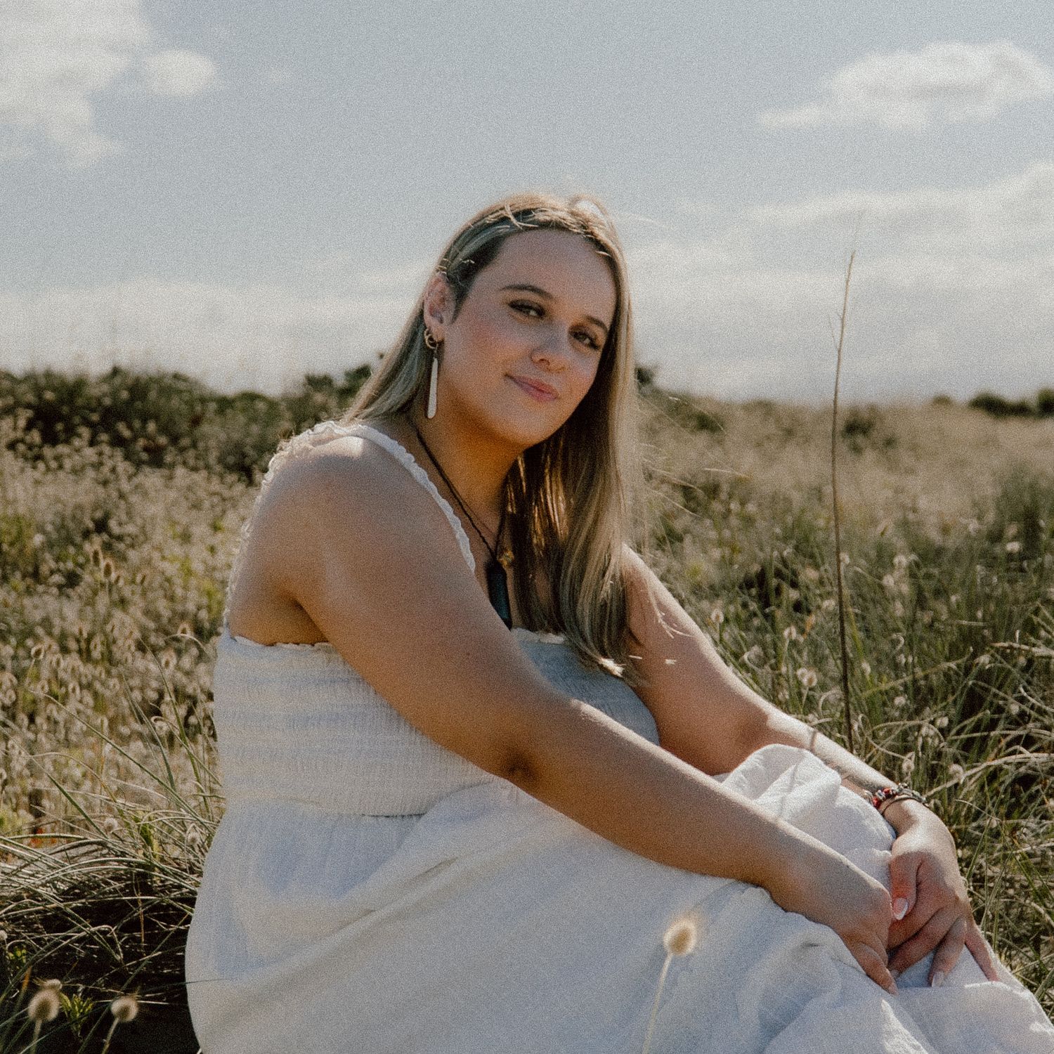 A person in a white dress sits thoughtfully in a sunlit meadow on a summer afternoon.