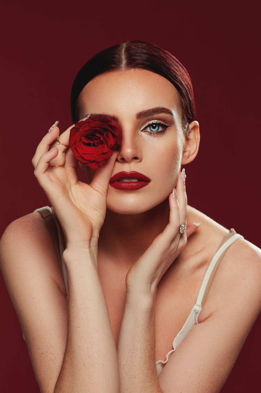 Artistic beauty portrait with red rose and dramatic makeup against dark burgundy background.