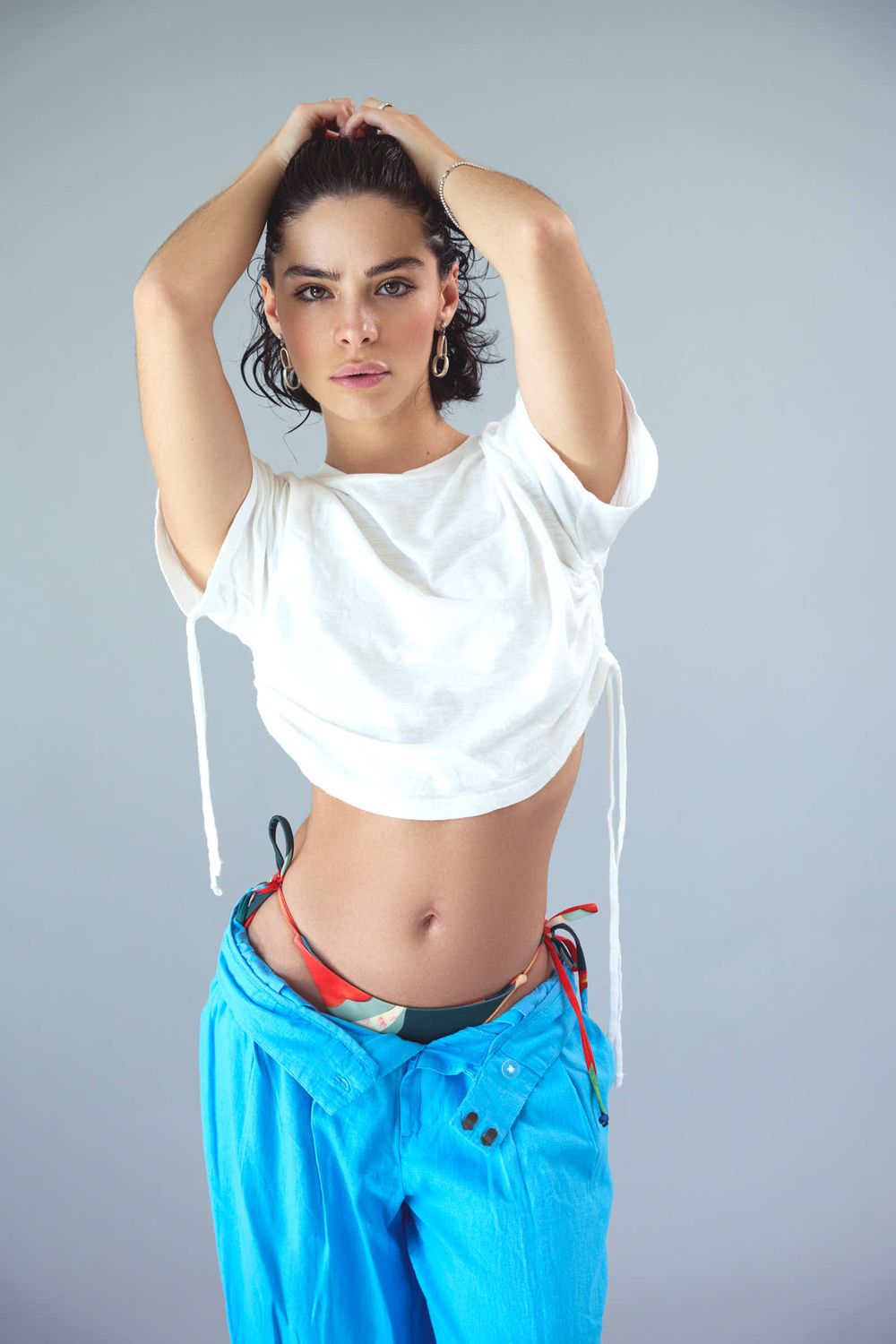 Athletic model in white crop top and bright blue pants strikes dynamic pose against gray backdrop.
