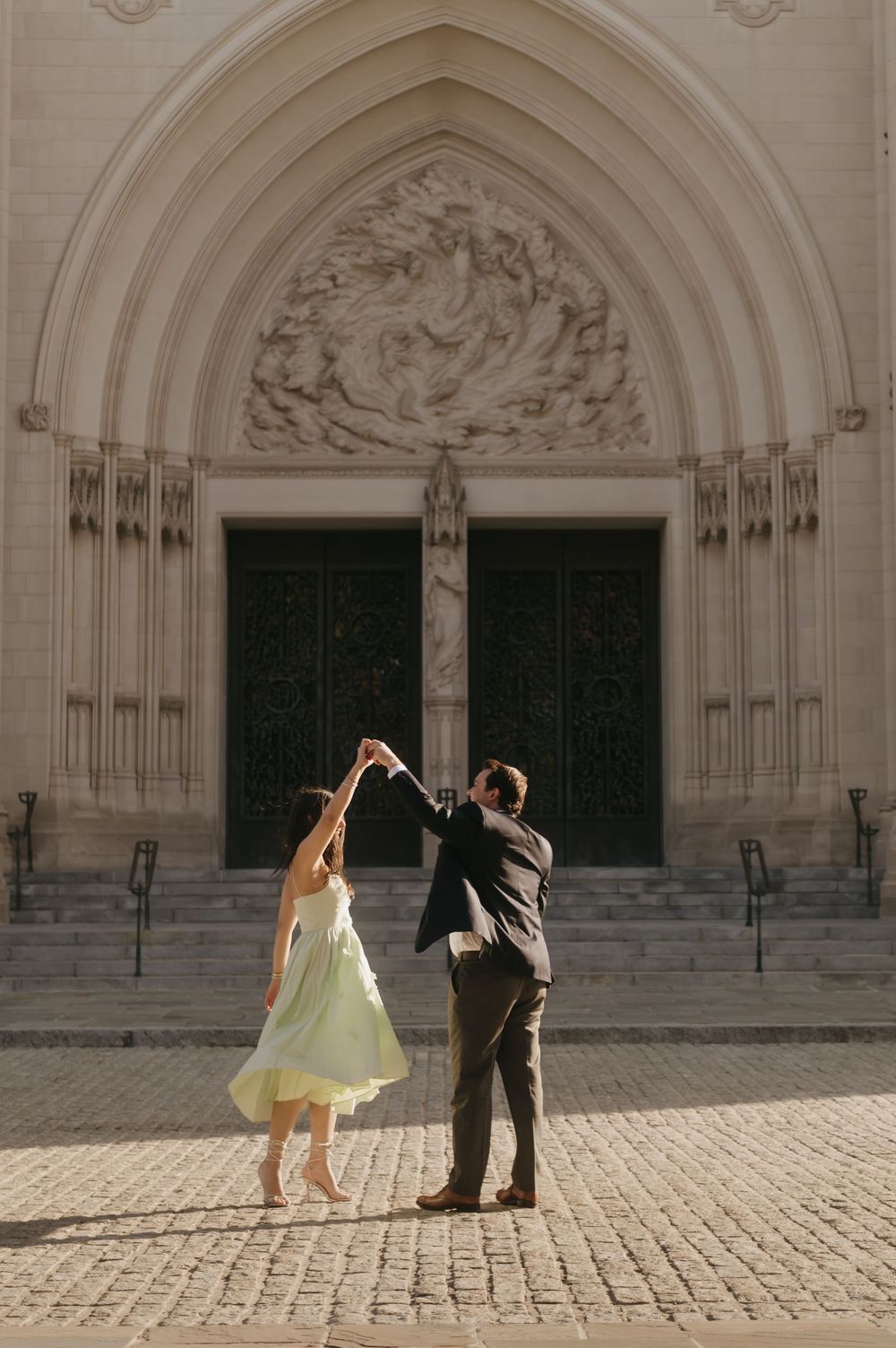 A couple in formal attire dances playfully in front of an ornate cathedral entrance with gothic arched doorways.