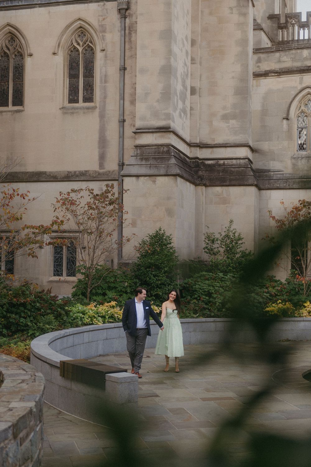 A couple strolls along a stone path beside a Gothic cathedral with autumn foliage.