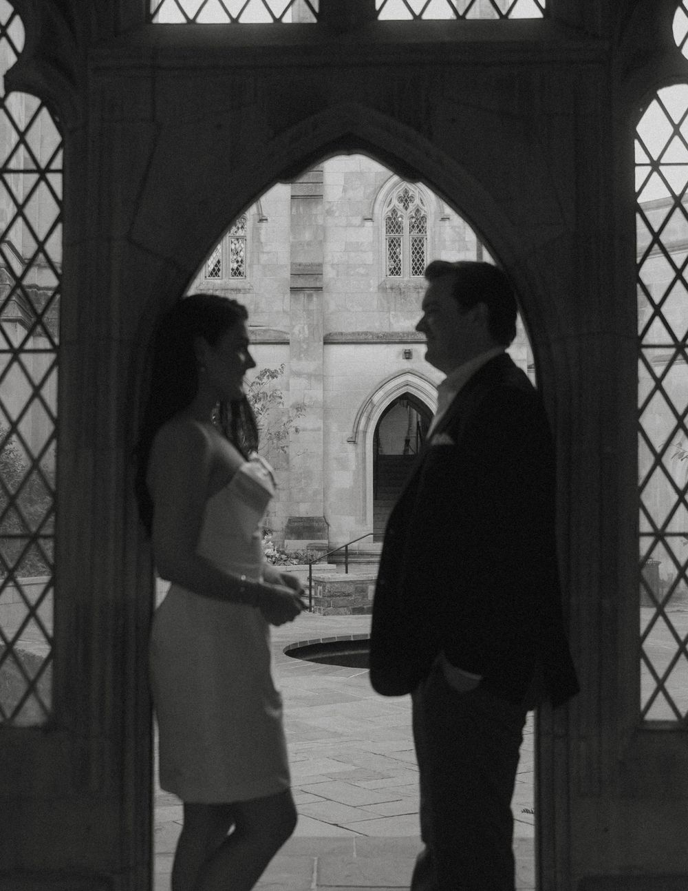 A black and white photo of a couple standing in a Gothic archway with diamond-paned windows.