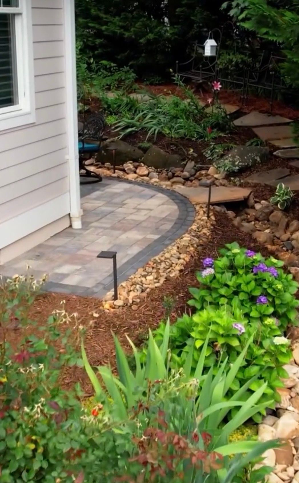 A curved paver patio with stone edging surrounded by lush green plants and purple flowers in a landscaped garden.