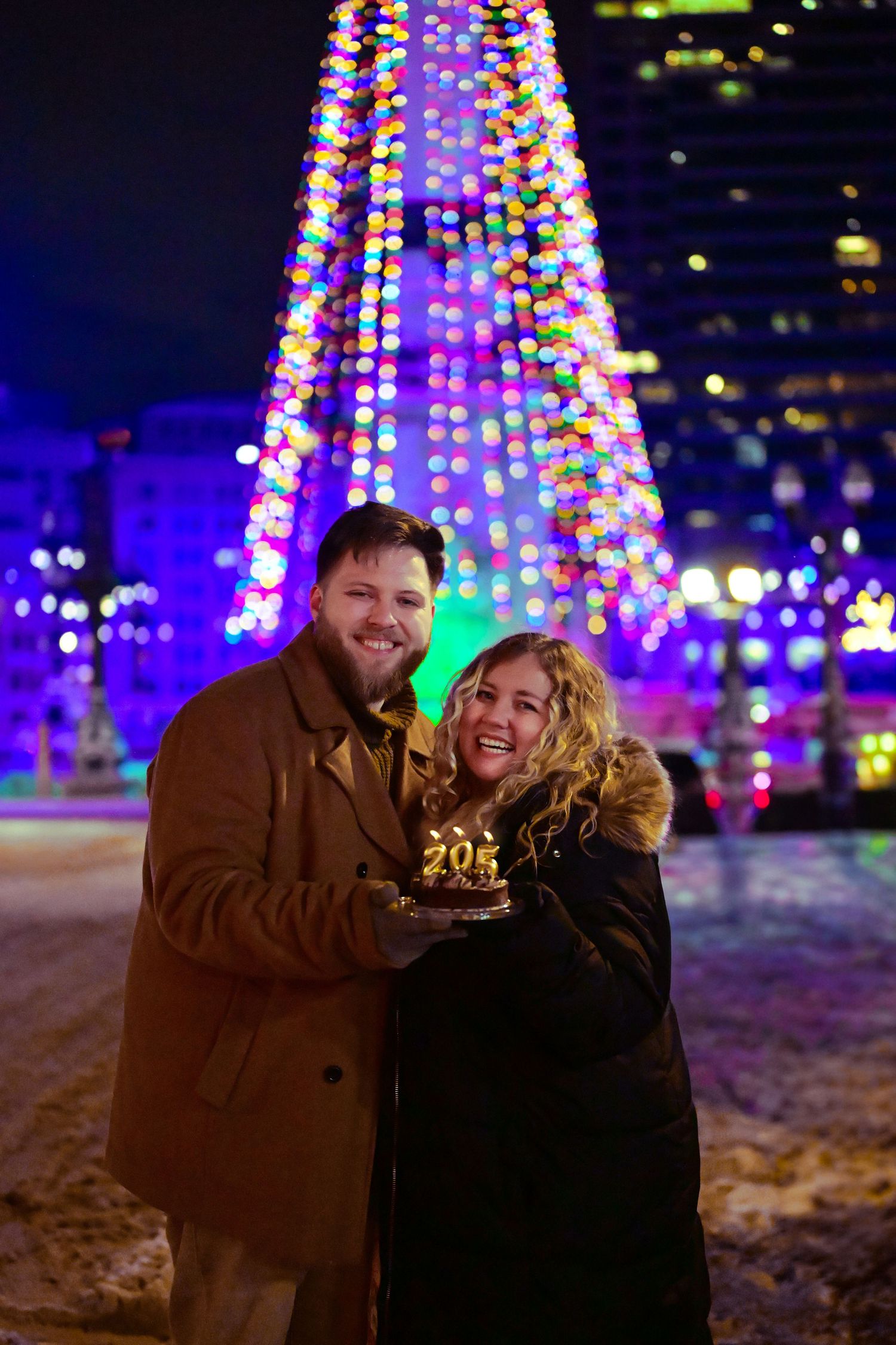 A couple celebrates with a 2024 sparkler in front of a tall illuminated Christmas tree display at night.