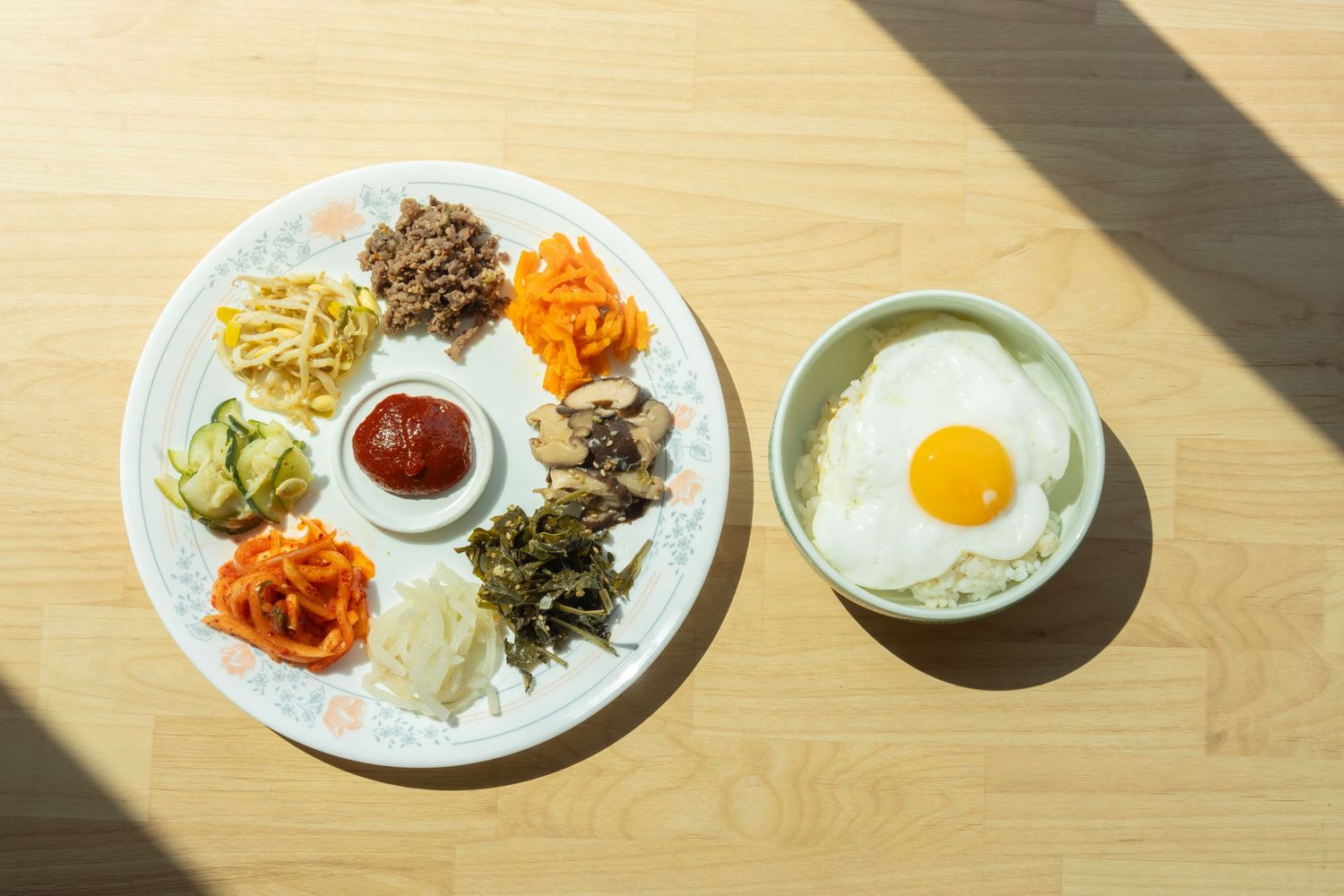 Korean bibimbap ingredients arranged on white plates with sunny side up egg and colorful vegetables in sunlight.