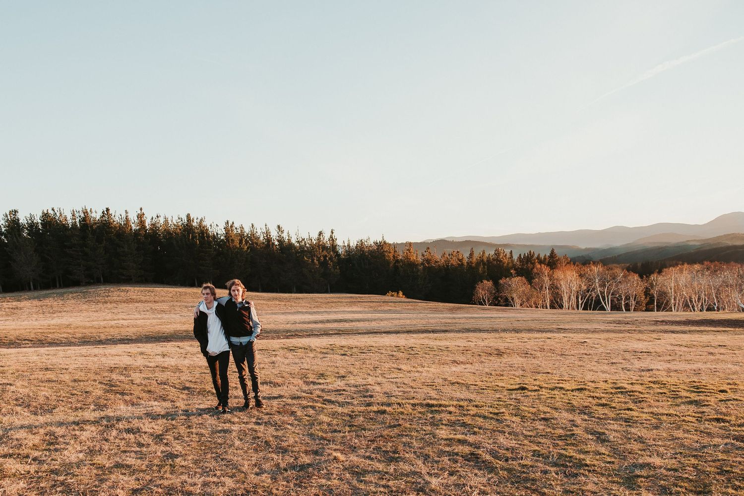 Family photographer Canberra Two people walk hand in hand through a golden field at sunset with mountains and forest in the distance.