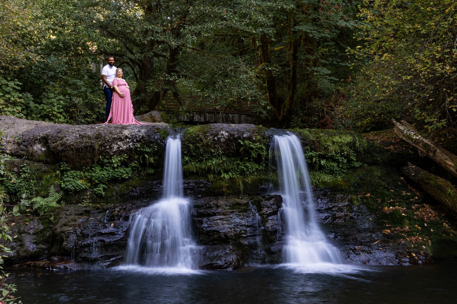 Couple embracing on rocks above a serene double waterfall surrounded by lush green forest trees.